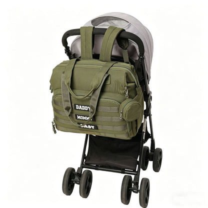 olive green diaper bag positioned on a stroller handle showing front webbing pocket dual handles and convertible carry design